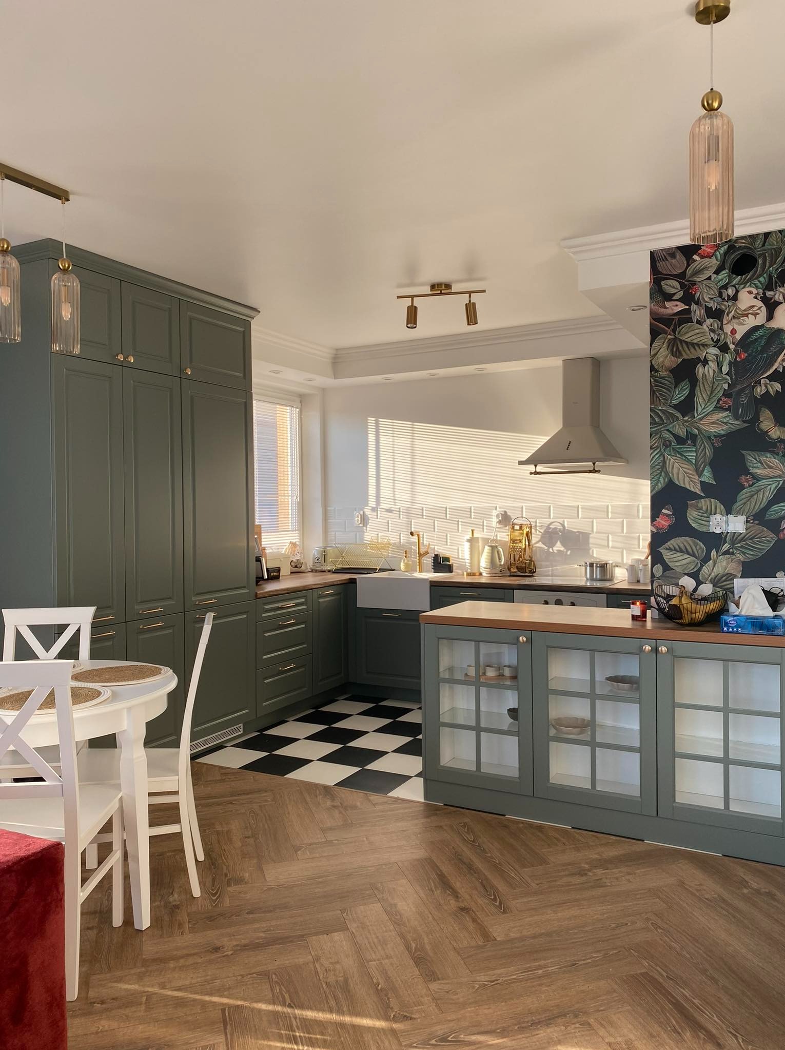 Nowoczesna kuchnia z zielonymi szafkami, białymi płytkami na ścianie i czarno-białą podłogą w szachownicę, drewniana podłoga ułożona w jodełkę, stół z białymi krzesłami i dekoracyjna tapeta...