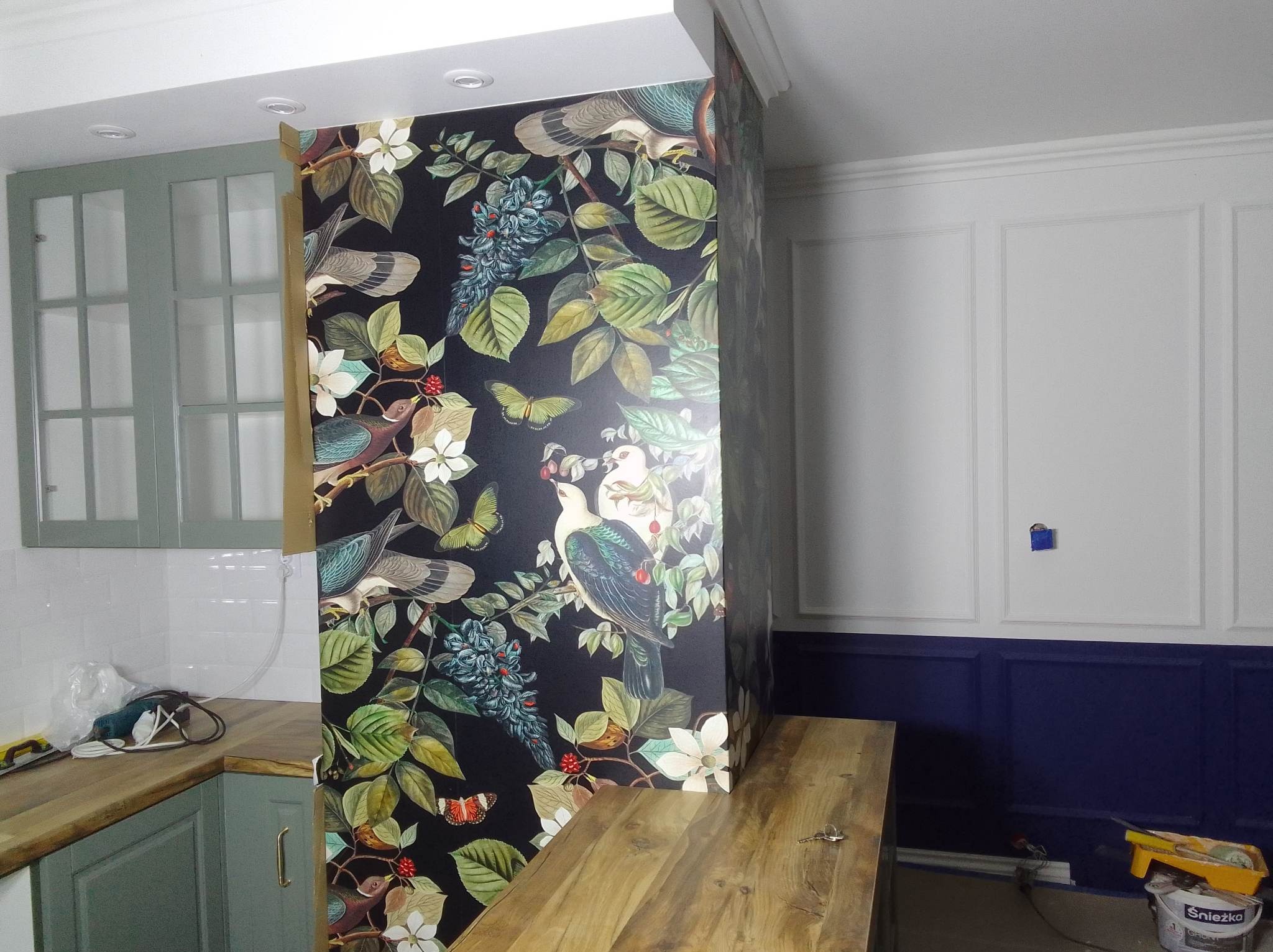 Fragment kuchni z wyspą obłożoną tapetą w motywy roślinne i ptasie, widoczne białe kafelki na ścianie, zielone szafki kuchenne oraz pomalowana na biało i granatowo ściana z dekoracyjnymi listwami.