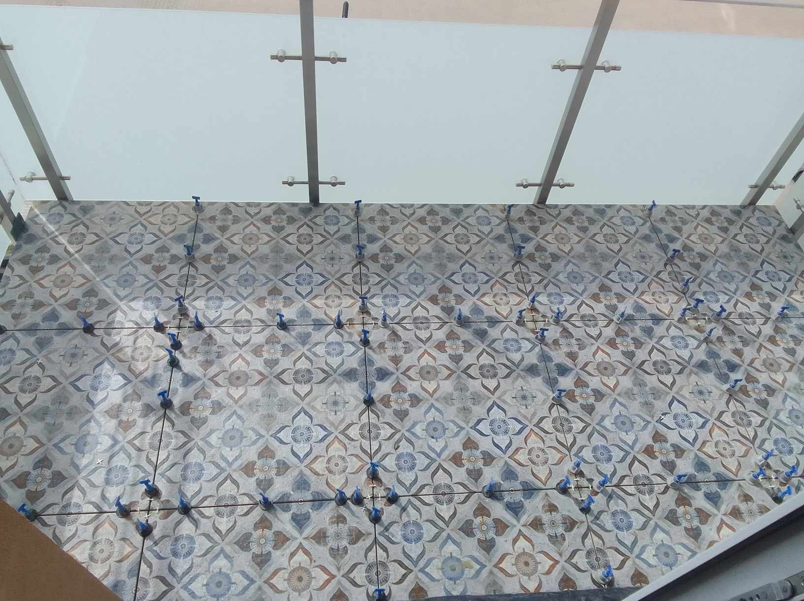 Ułożone płytki pod szklanym zadaszeniem, z niebieskimi klinami poziomującymi w fugach, wzór płytek w odcieniach szarości, brązu i błękitu.