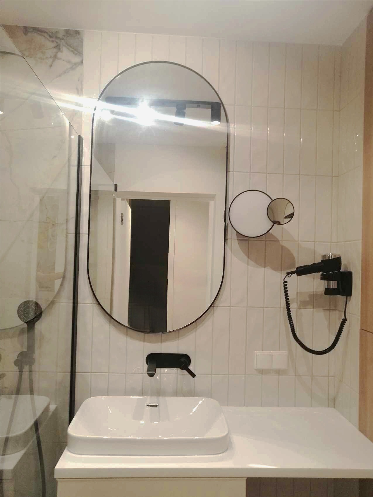 Wykończona łazienka z owalnym lustrem w czarnej ramie, białą umywalką nablatową, czarną armaturą i suszarką do włosów na ścianie z białych, pionowo ułożonych płytek.