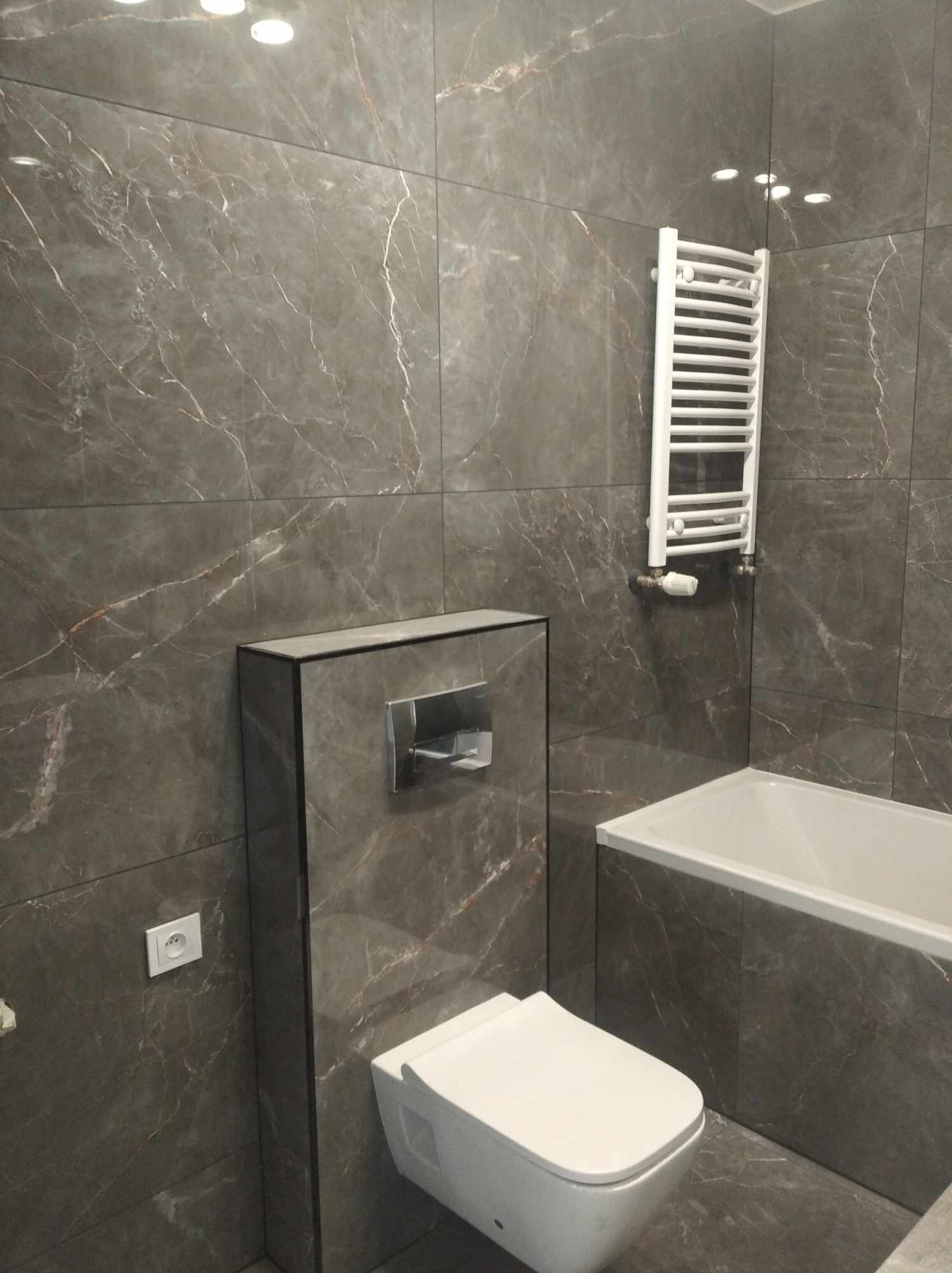 Nowoczesna łazienka z szarymi płytkami imitującymi marmur, biała armatura (toaleta, wanna) i grzejnik drabinkowy, oświetlenie punktowe w suficie.