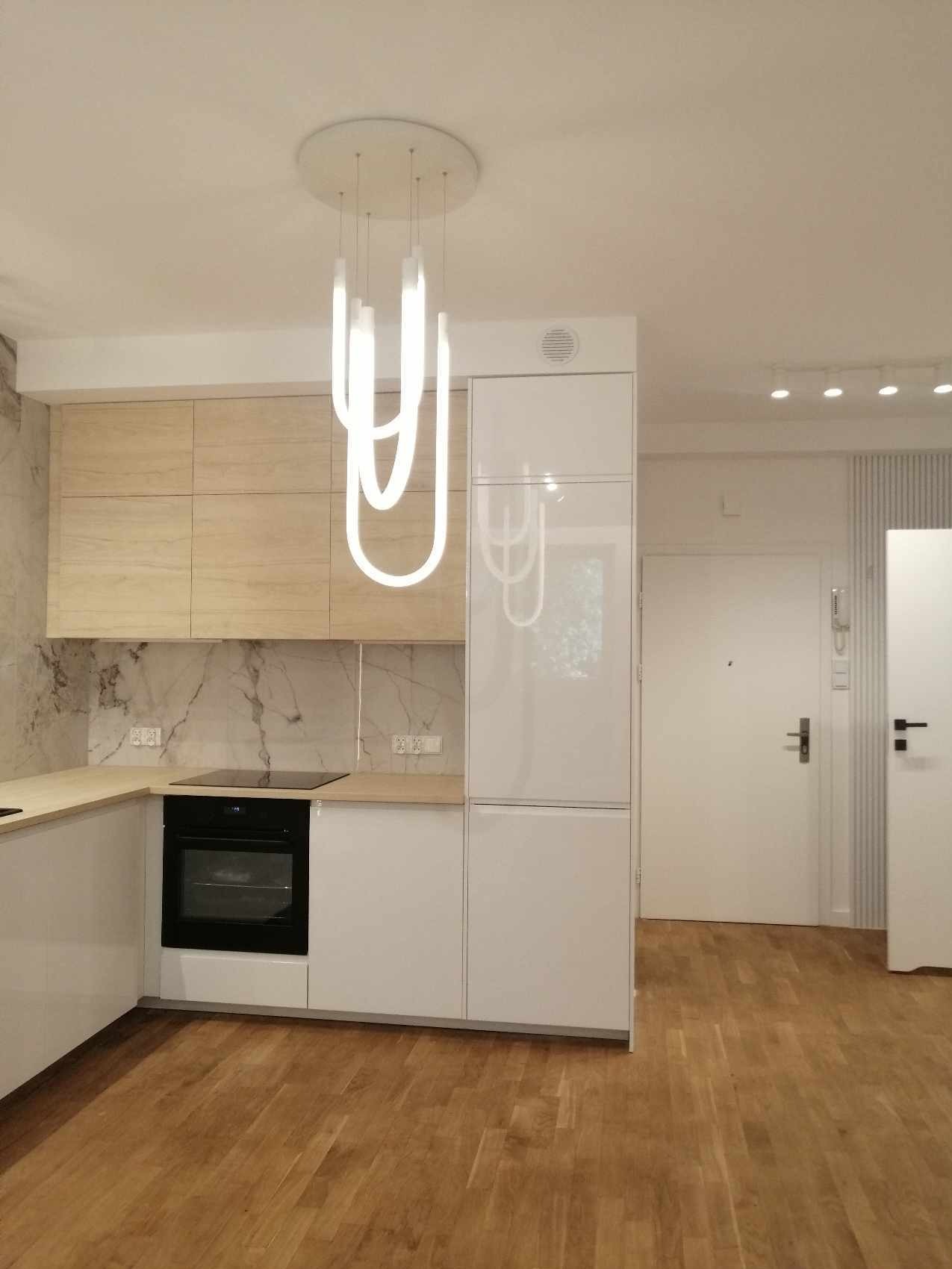 Nowoczesna kuchnia z białymi szafkami, drewnianymi blatami i marmurowym backsplashiem, oświetlona designerską lampą wiszącą z ledowych rurek.