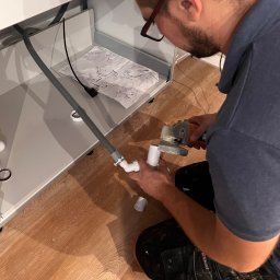 ReZmont BARTŁOMIEJ REZNEROWICZ - Instalator z brodą w okularach, przycina rurę PCV szlifierką kątową pod umywalką w łazience, widoczne instrukcje montażu na blacie szafki.