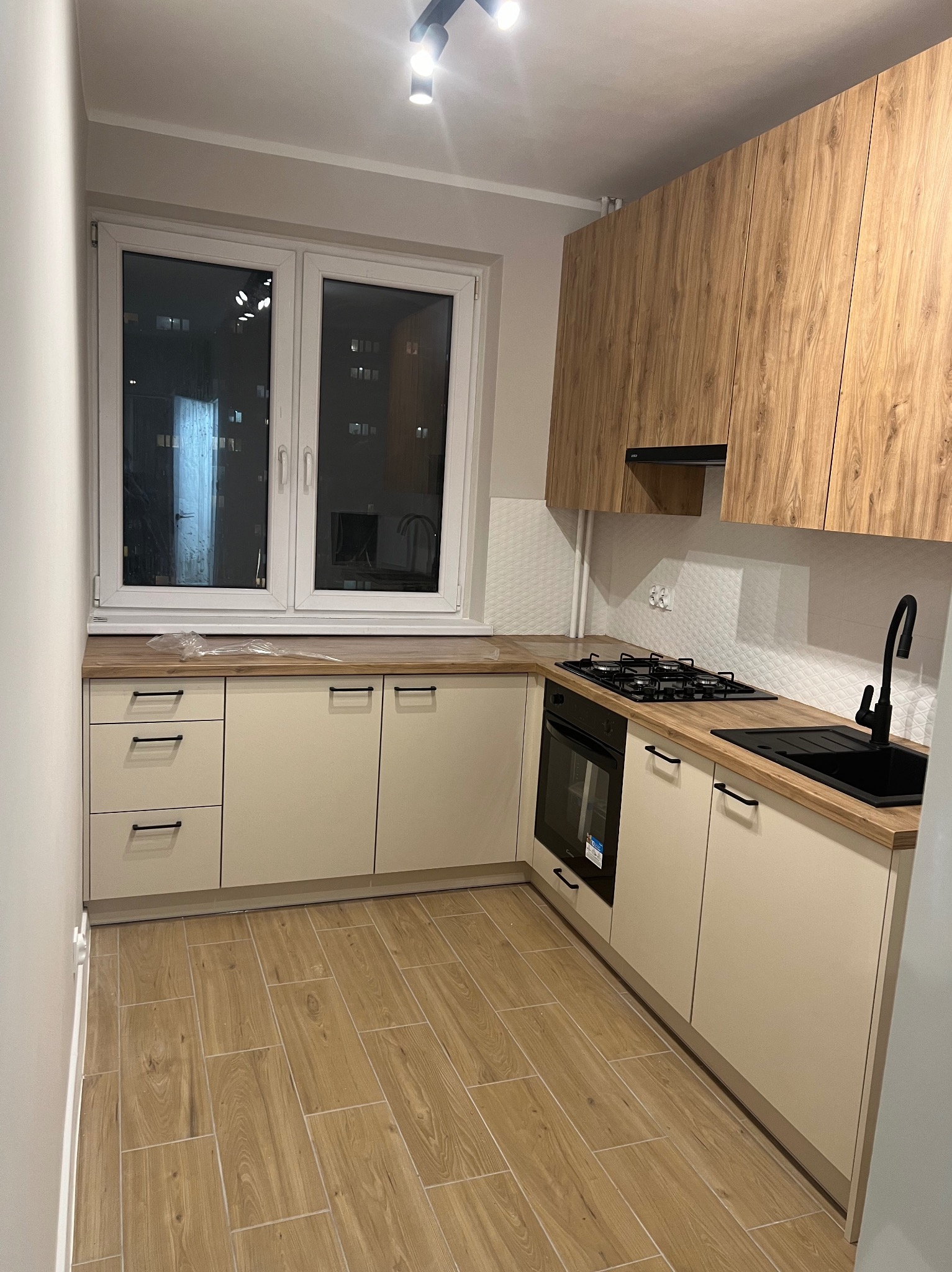 Nowoczesna kuchnia z beżowymi szafkami dolnymi i drewnopodobnymi górnymi, czarną armaturą i płytą grzewczą, wykończona drewnopodobnymi płytkami podłogowymi i białymi płytkami ściennymi.