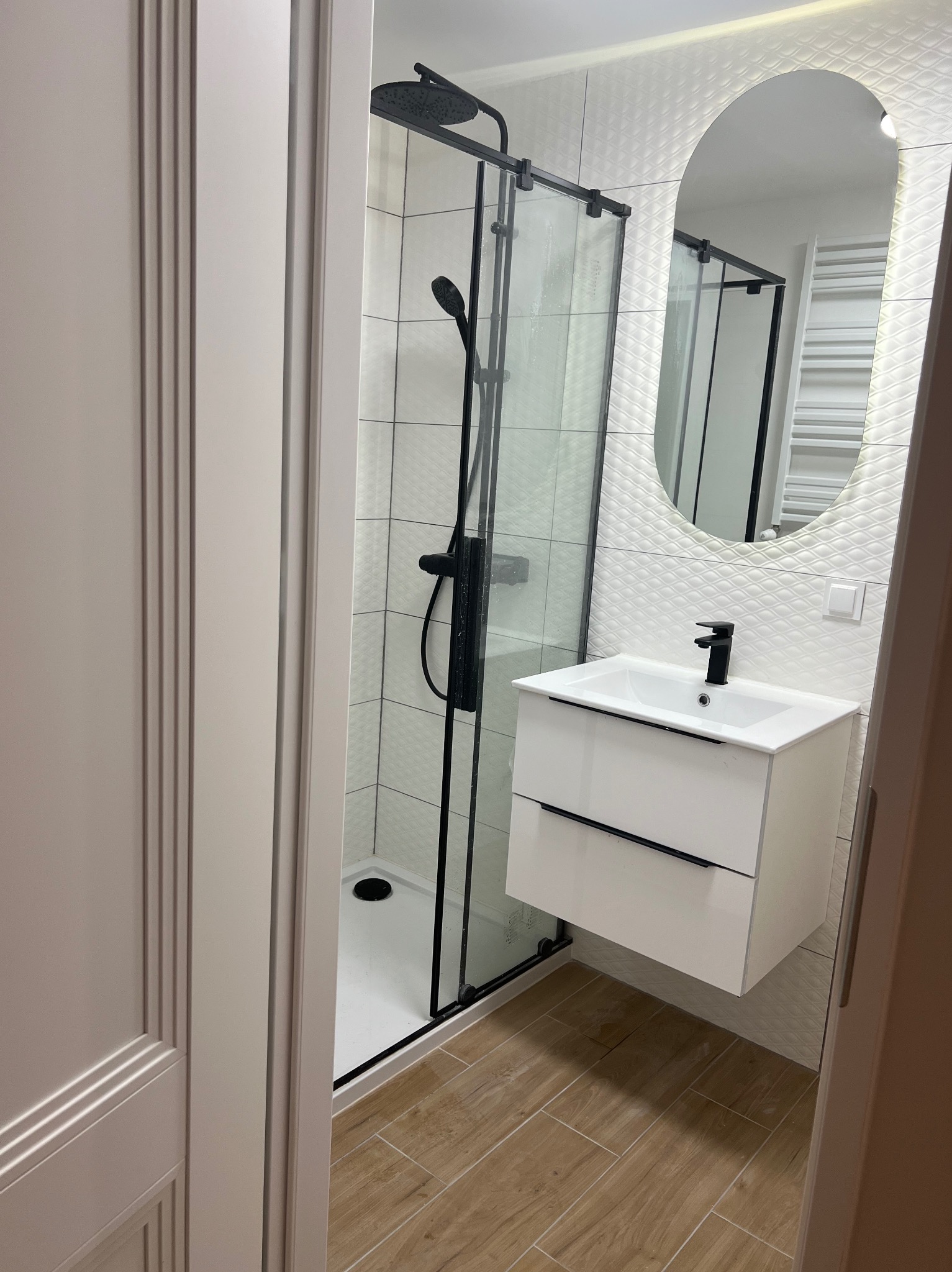 Nowoczesna łazienka z kabiną prysznicową typu walk-in z czarnymi detalami, białą szafką pod umywalkę z czarnym uchwytem, owalnym lustrem z podświetleniem LED i płytkami 3D na ścianie.