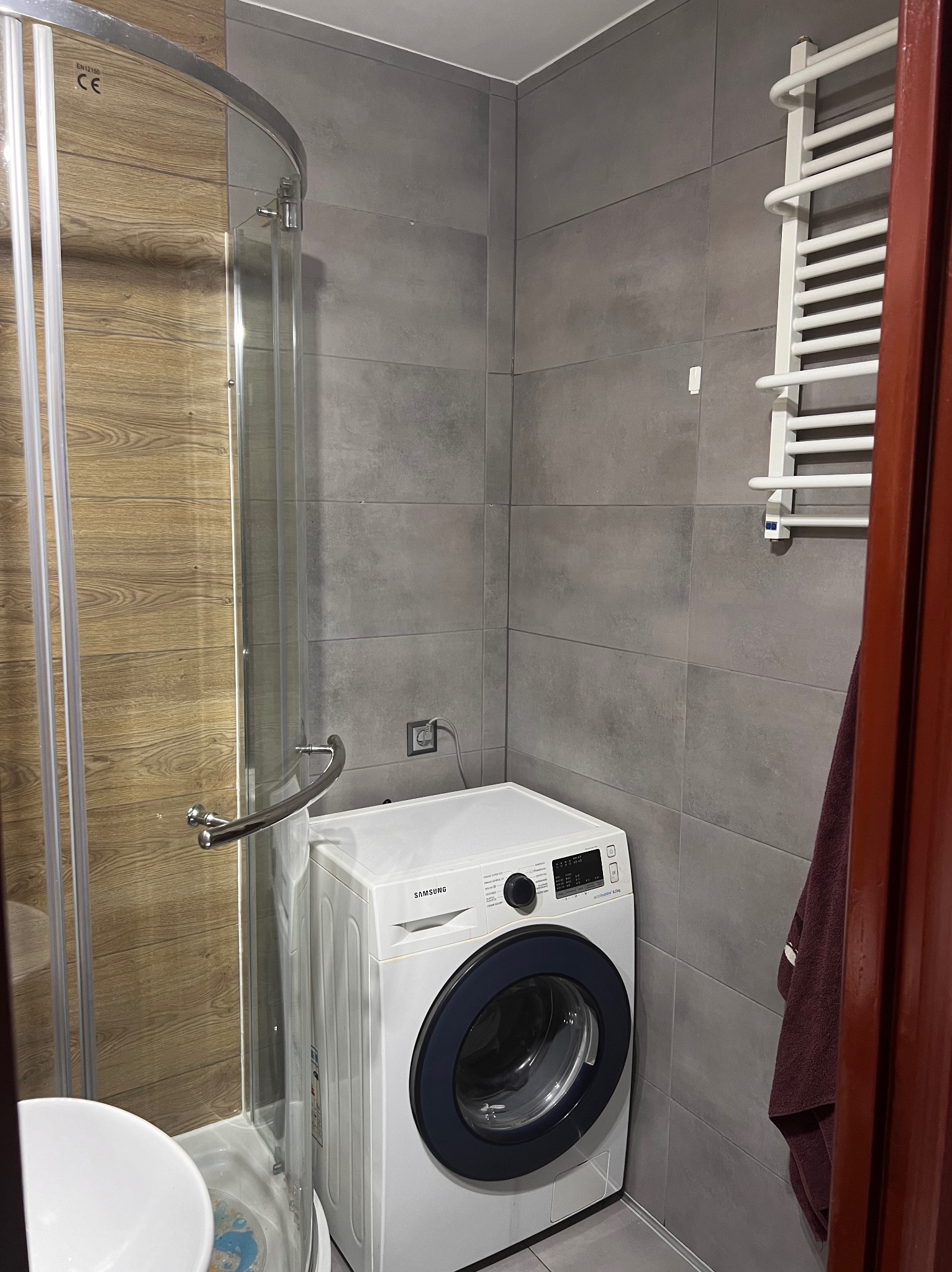 Szara łazienka z kabiną prysznicową z drewnopodobnymi panelami, białą pralką Samsung i grzejnikiem drabinkowym.