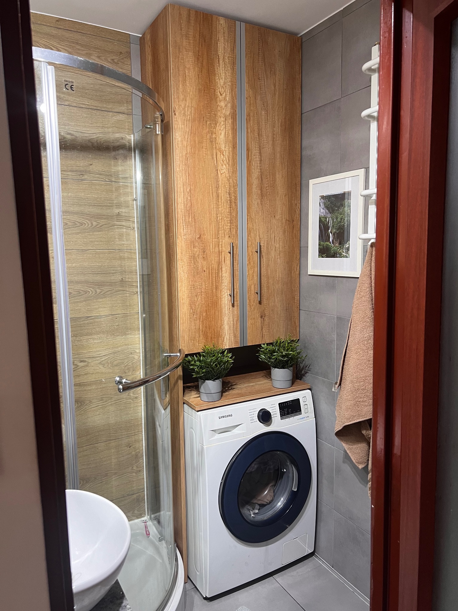 Nowoczesna łazienka z kabiną prysznicową, pralką Samsung pod drewnianym blatem z dekoracyjnymi roślinami w doniczkach, szare płytki na ścianach i drewnopodobne wykończenie kabiny prysznicowej.