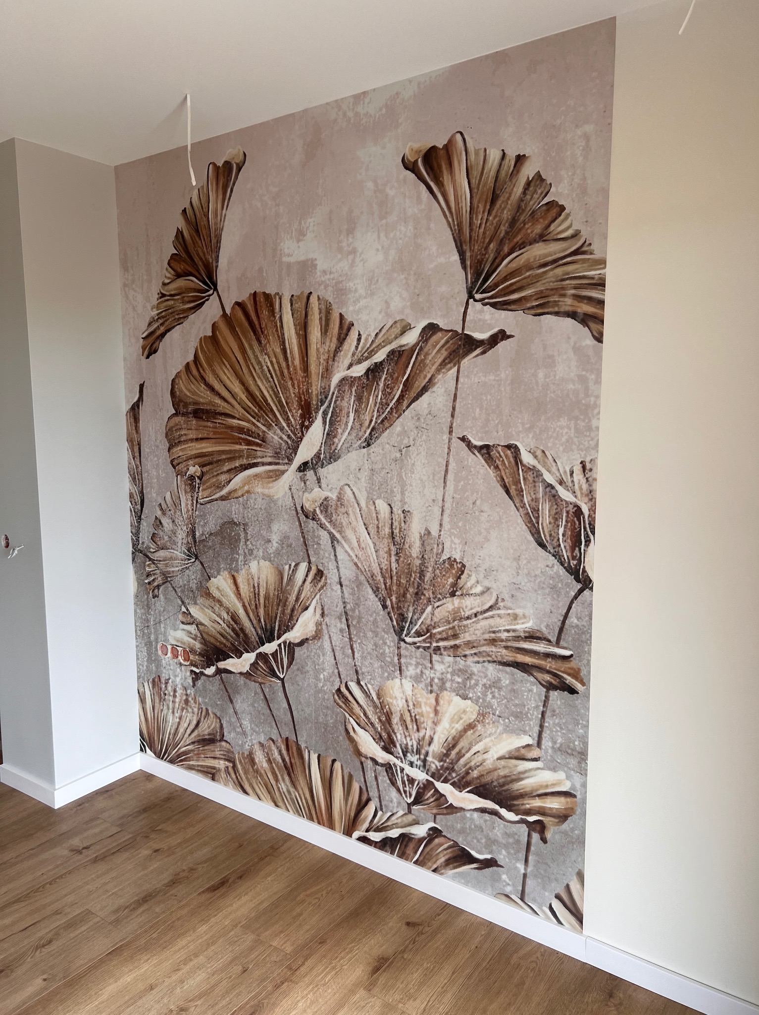 Ściana z fototapetą przedstawiającą brązowe liście na tle imitującym betonową ścianę, widoczne drewniane panele podłogowe i biała listwa przypodłogowa.