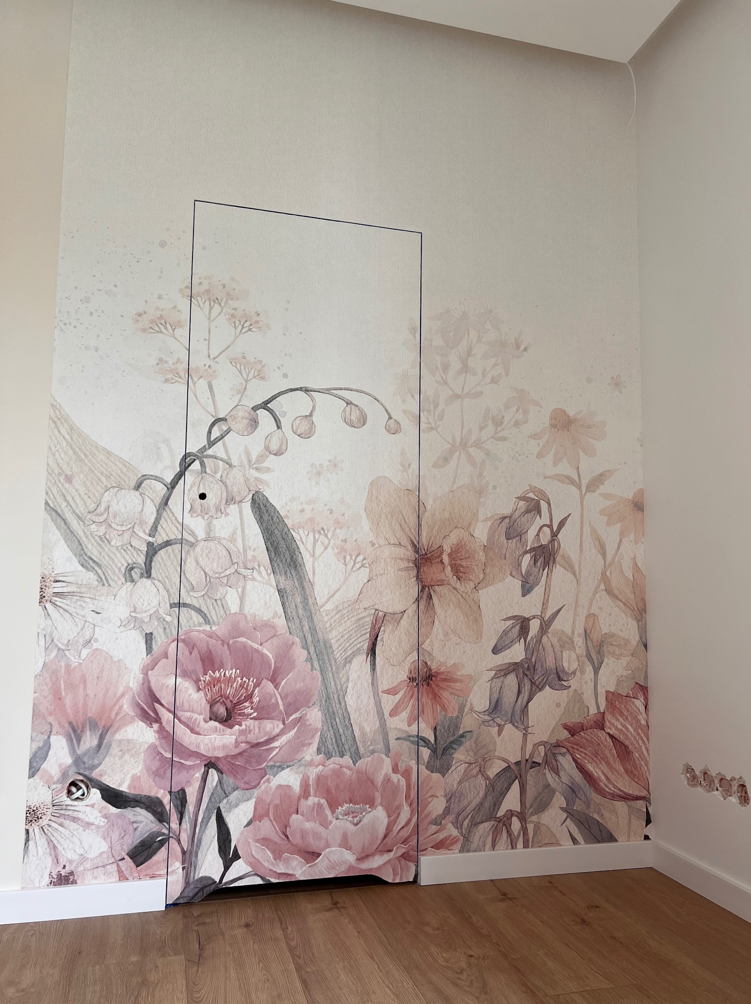 Ściana w pokoju z tapetą w pastelowe kwiaty, imitująca ukryte drzwi, widoczne gniazdka elektryczne w ścianie oraz drewniana podłoga.