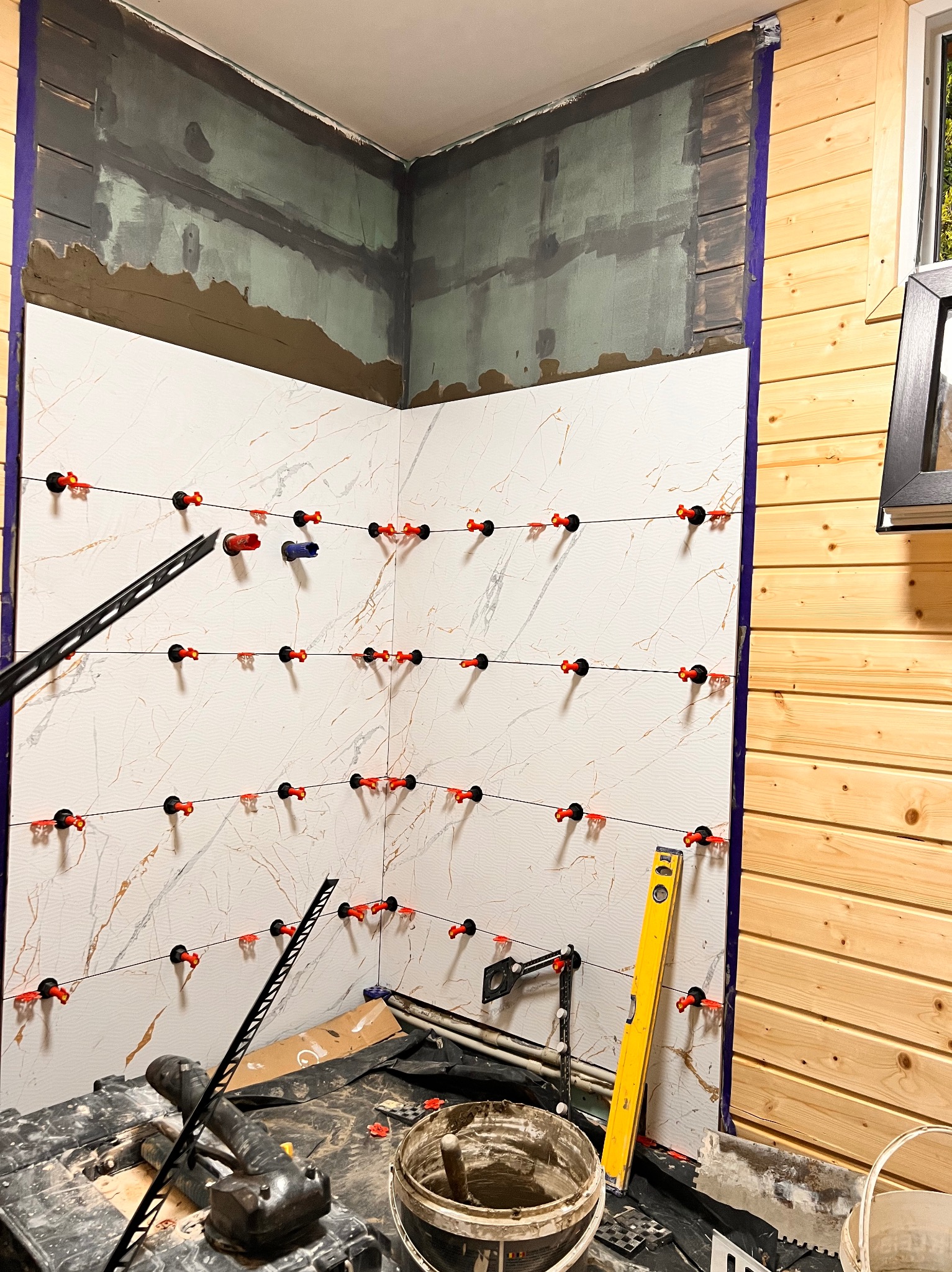 Ściana w trakcie układania płytek imitujących marmur, z użyciem systemu poziomowania płytek, widoczny poziomica i przygotowane podłoże na górze ściany.