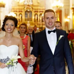 Uśmiechnięta para młoda, panna młoda w białej sukni z kaliami i welonem, pan młody w granatowym garniturze z muszką, trzymają się za ręce w kościele z pozłacanym ołtarzem w tle.