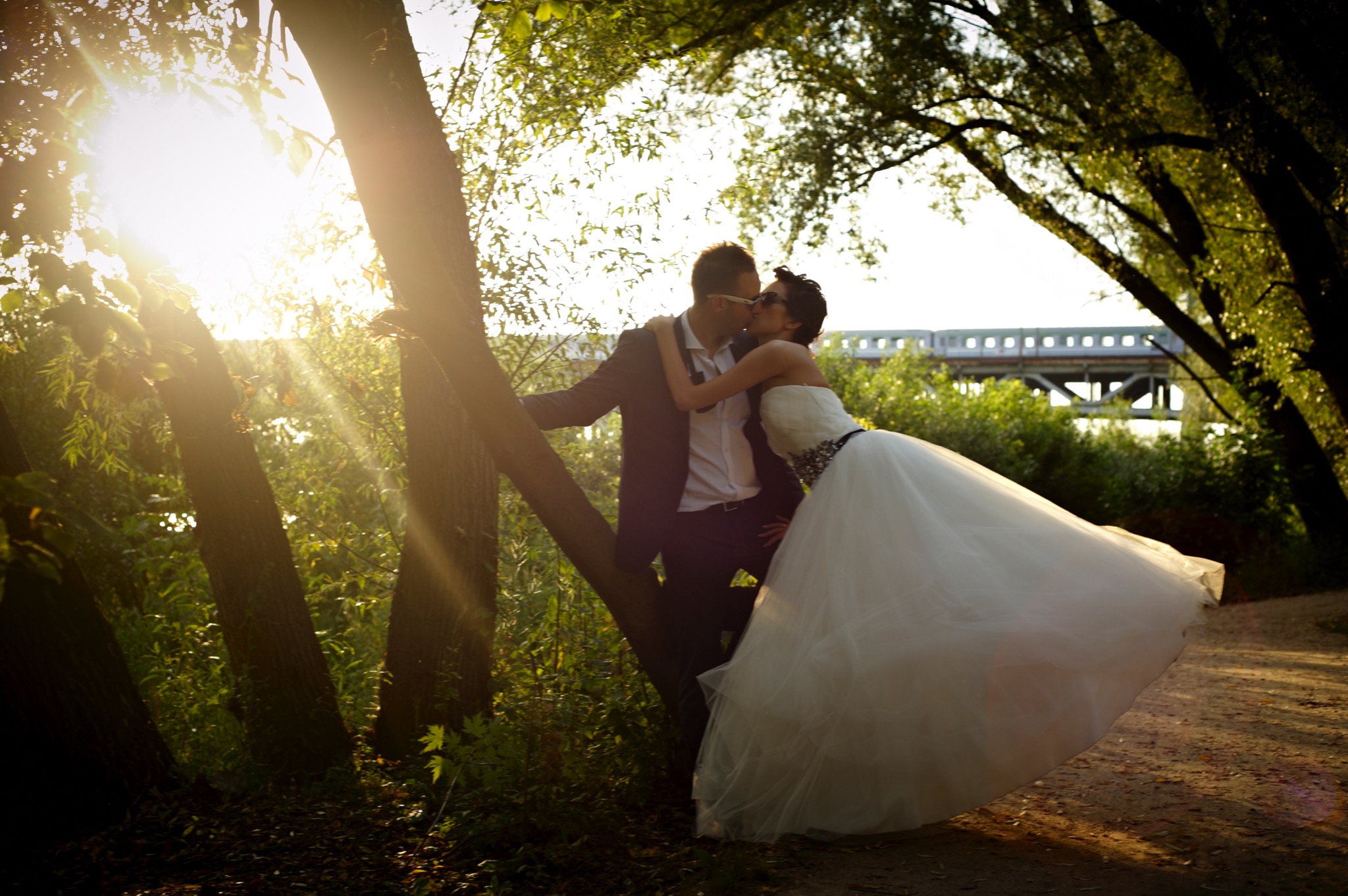 Para młoda całuje się opierając o drzewo, w tle widoczny most kolejowy, promienie słońca przebijają przez liście.