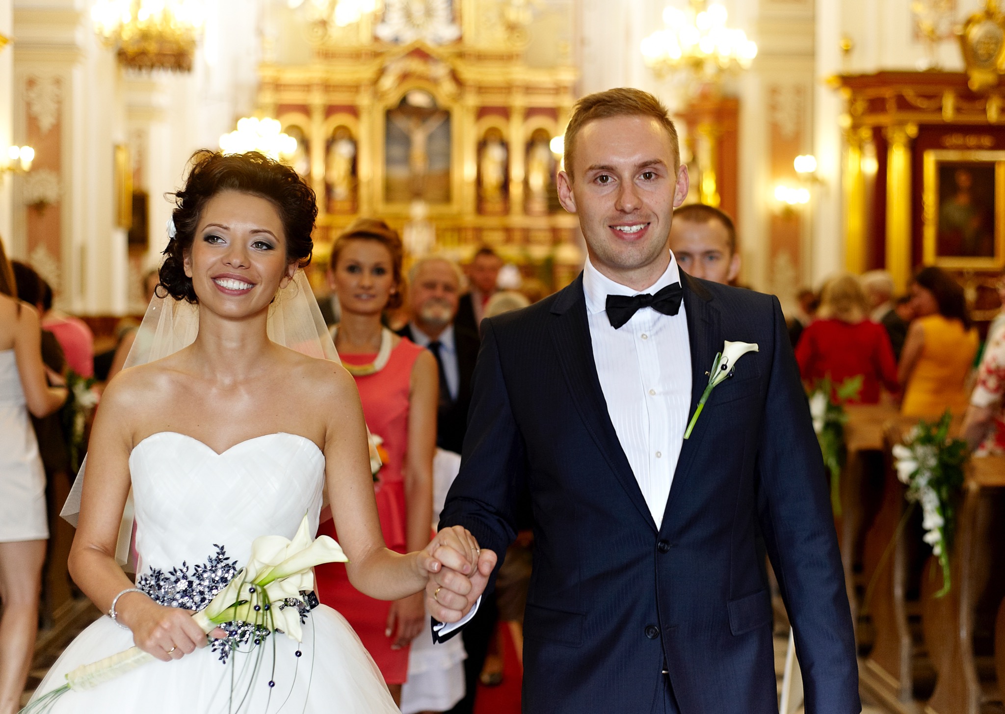 Uśmiechnięta para młoda, panna młoda w białej sukni z kaliami i welonem, pan młody w granatowym garniturze z muszką, trzymają się za ręce w kościele z pozłacanym ołtarzem w tle.