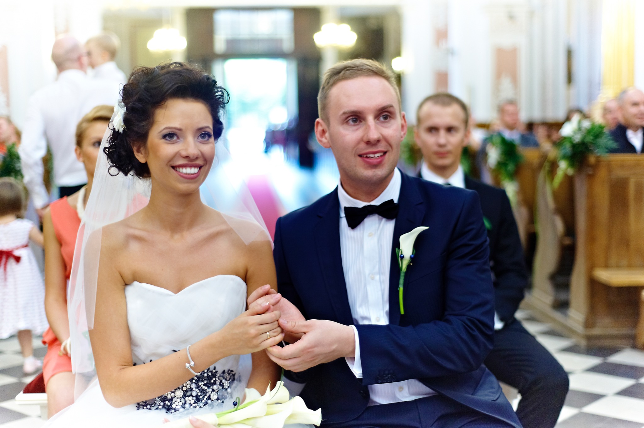 Para młoda siedzi w kościele, trzymając się za ręce, z gośćmi weselnymi w tle i bukietem białych kwiatów.
