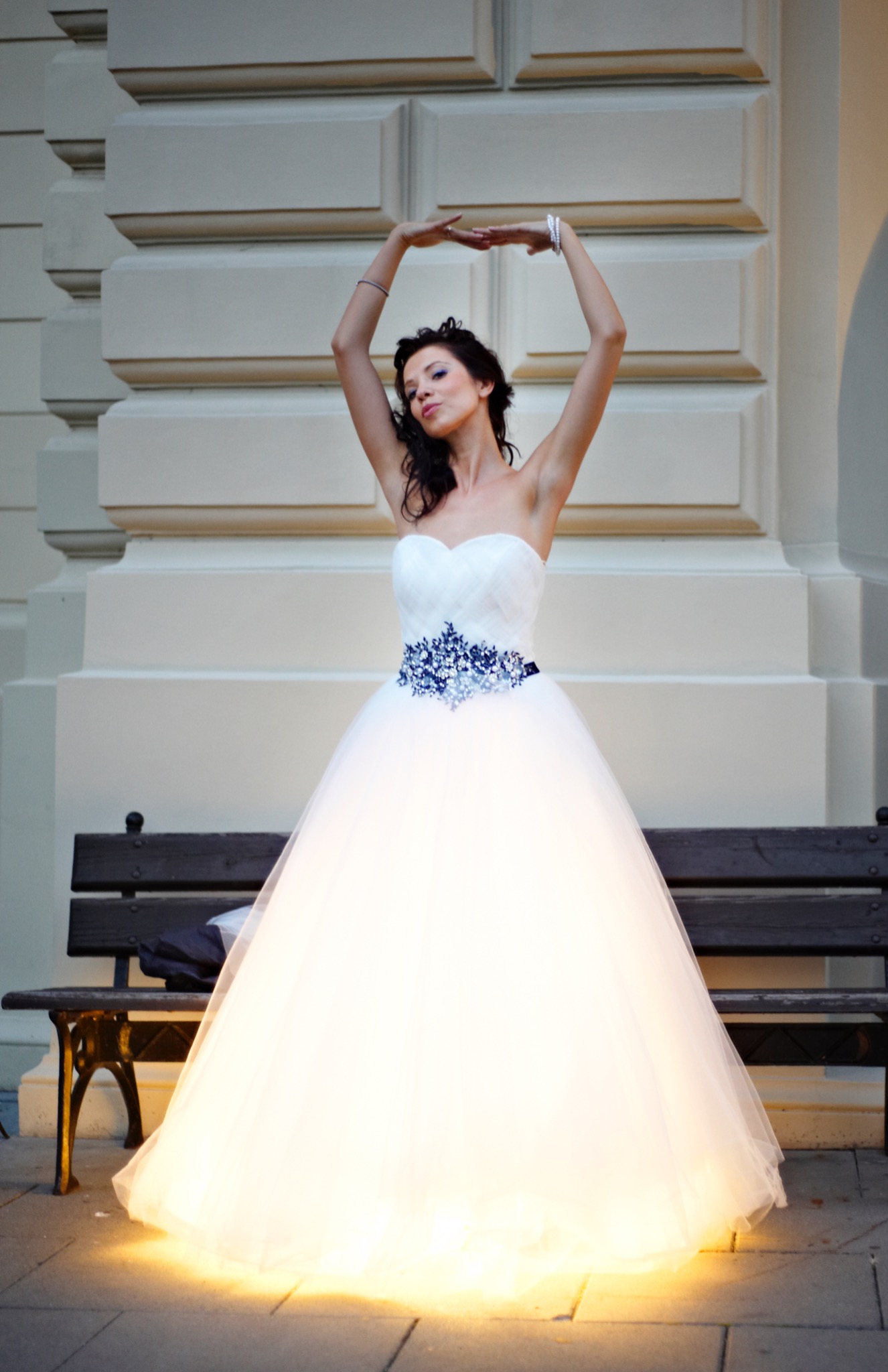 Panna młoda w białej sukni balowej z niebieskim pasem, pozująca z uniesionymi rękami na tle klasycznej architektury i ławki.