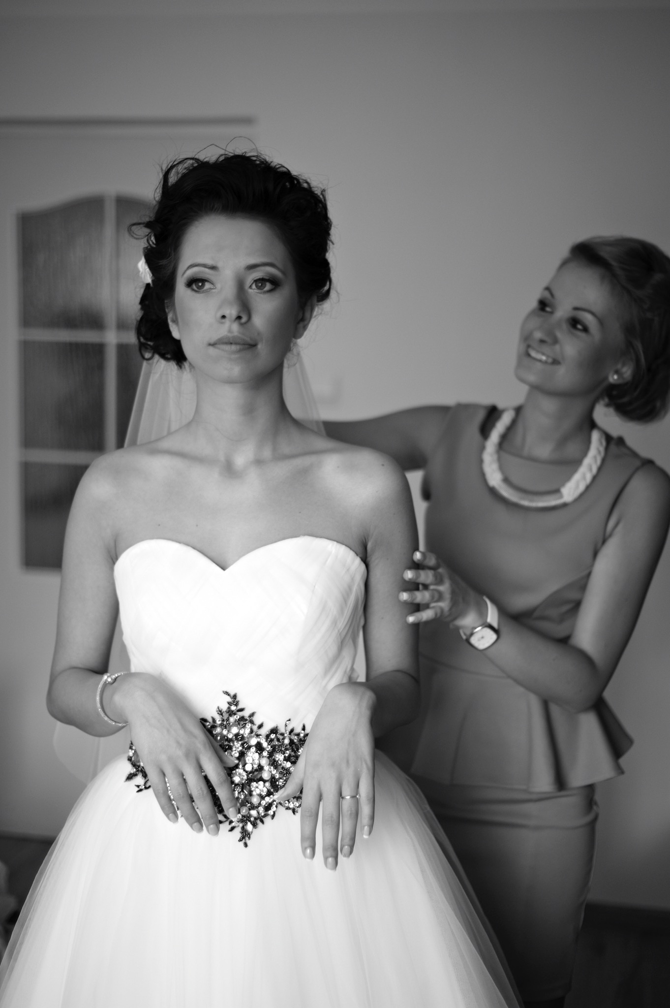 Panna młoda w sukni ślubnej z ozdobnym czarnym pasem, poprawiana przez druhnę, czarno-białe zdjęcie.