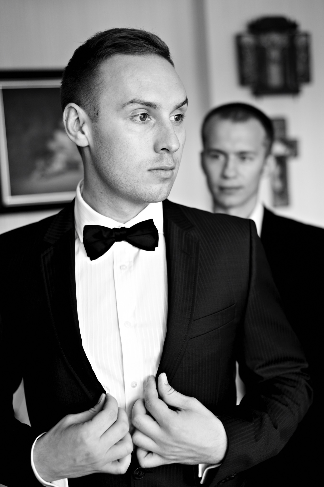 Czarno-białe zdjęcie pana młodego w garniturze z muchą, zapinającego marynarkę, z drugim mężczyzną w tle.