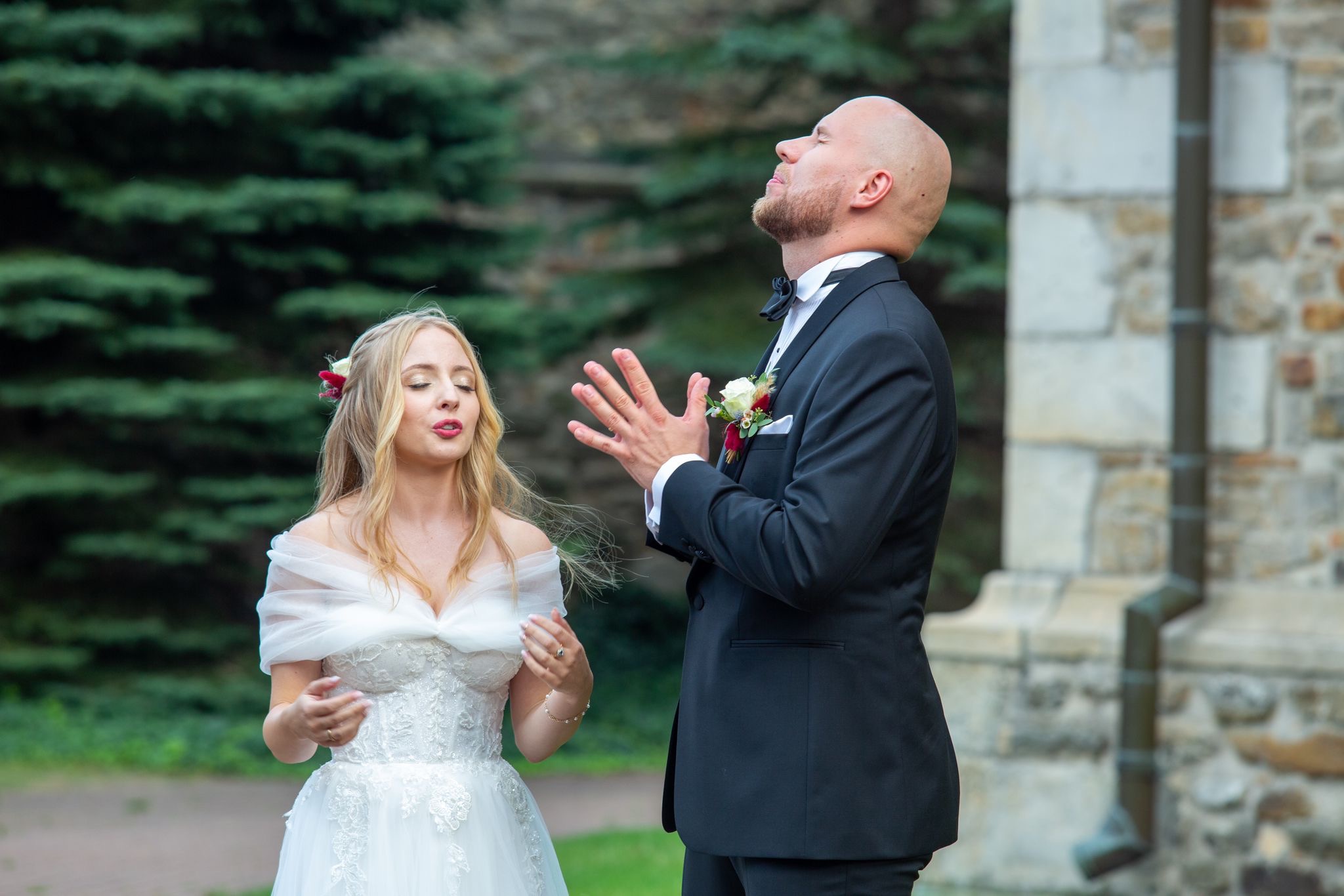 Panna młoda w białej sukni z kwiatami we włosach i pan młody w czarnym smokingu z zamkniętymi oczami i uniesionymi dłońmi, stojący przed kamiennym budynkiem i zielenią.