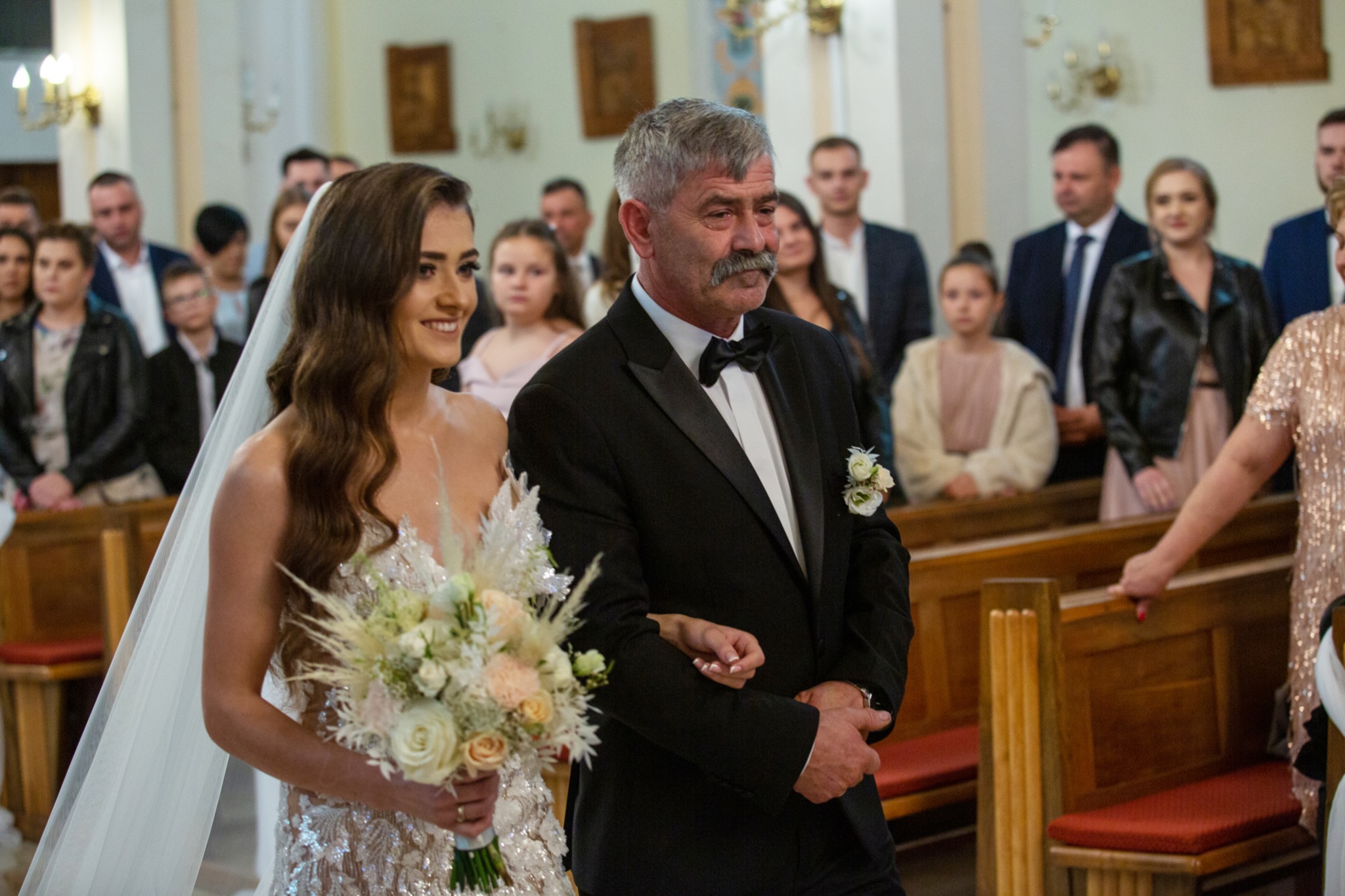 Panna młoda w sukni z cekinami, trzymająca bukiet z białych i kremowych róż, prowadzona do ołtarza przez ojca w czarnym smokingu na tle gości siedzących w ławkach kościoła.