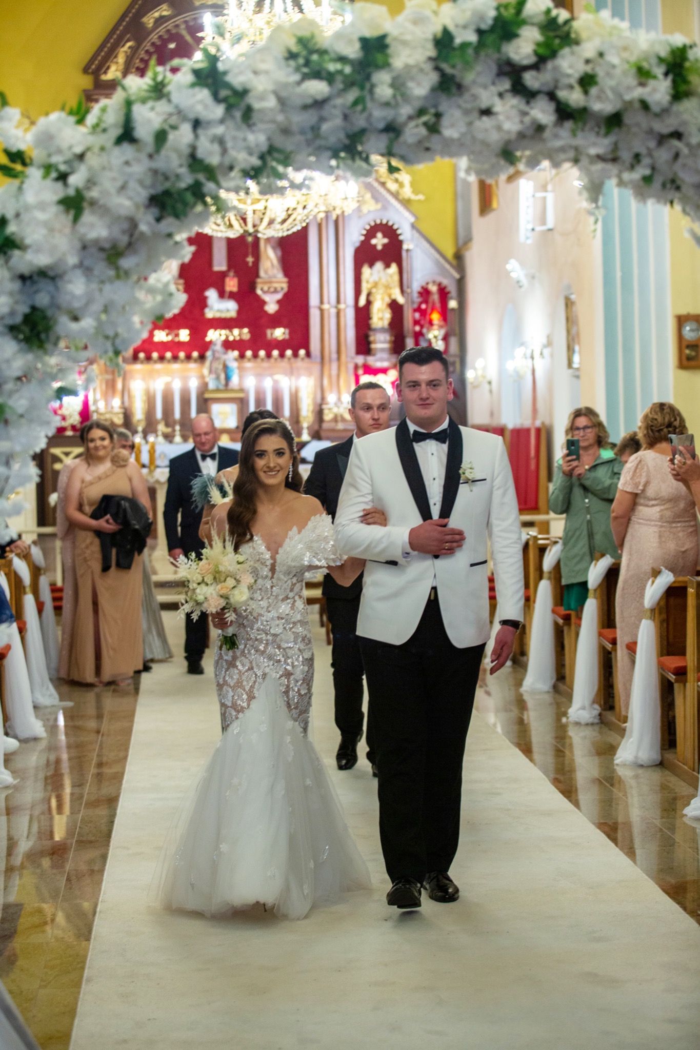 Para młoda idzie do ołtarza w kościele, panna młoda w koronkowej sukni z bukietem, pan młody w białym smokingu, dekoracja kwiatowa nad przejściem.