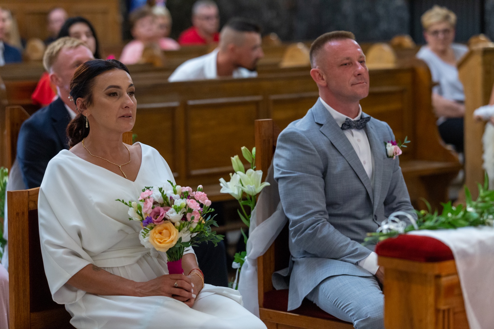 Para młoda siedzi w ławce kościelnej podczas ceremonii, panna młoda trzyma bukiet z różową wstążką, pan młody w szarym garniturze z muchą.