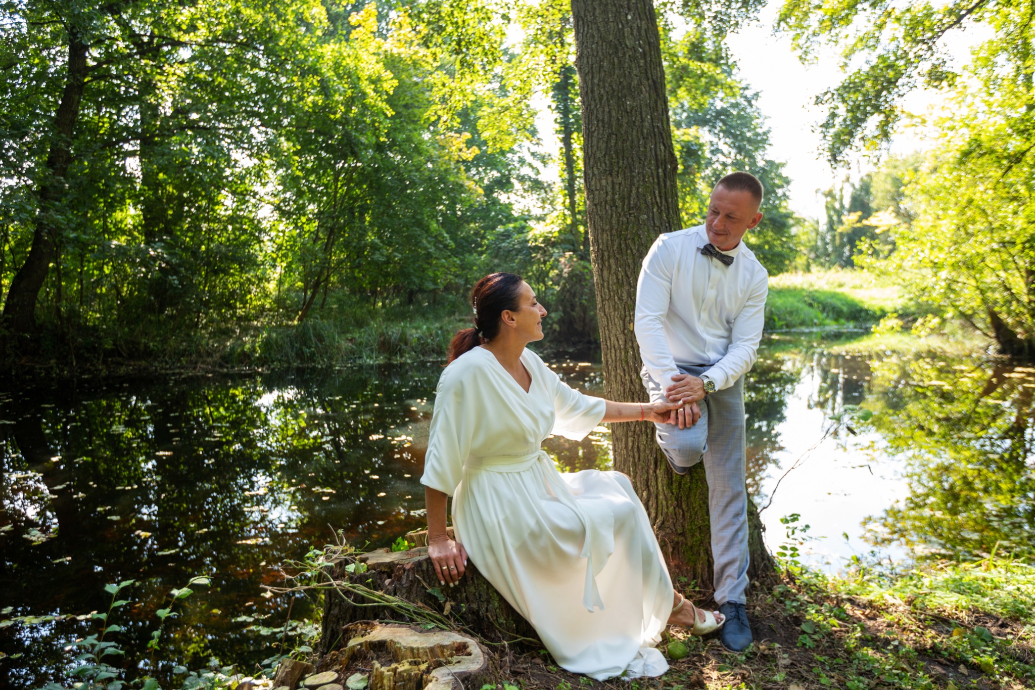 Para w eleganckich strojach pozuje na tle jeziora w lesie. Kobieta w białej sukni siedzi na pniu drzewa, mężczyzna w koszuli z muszką stoi obok, opierając się o drzewo.