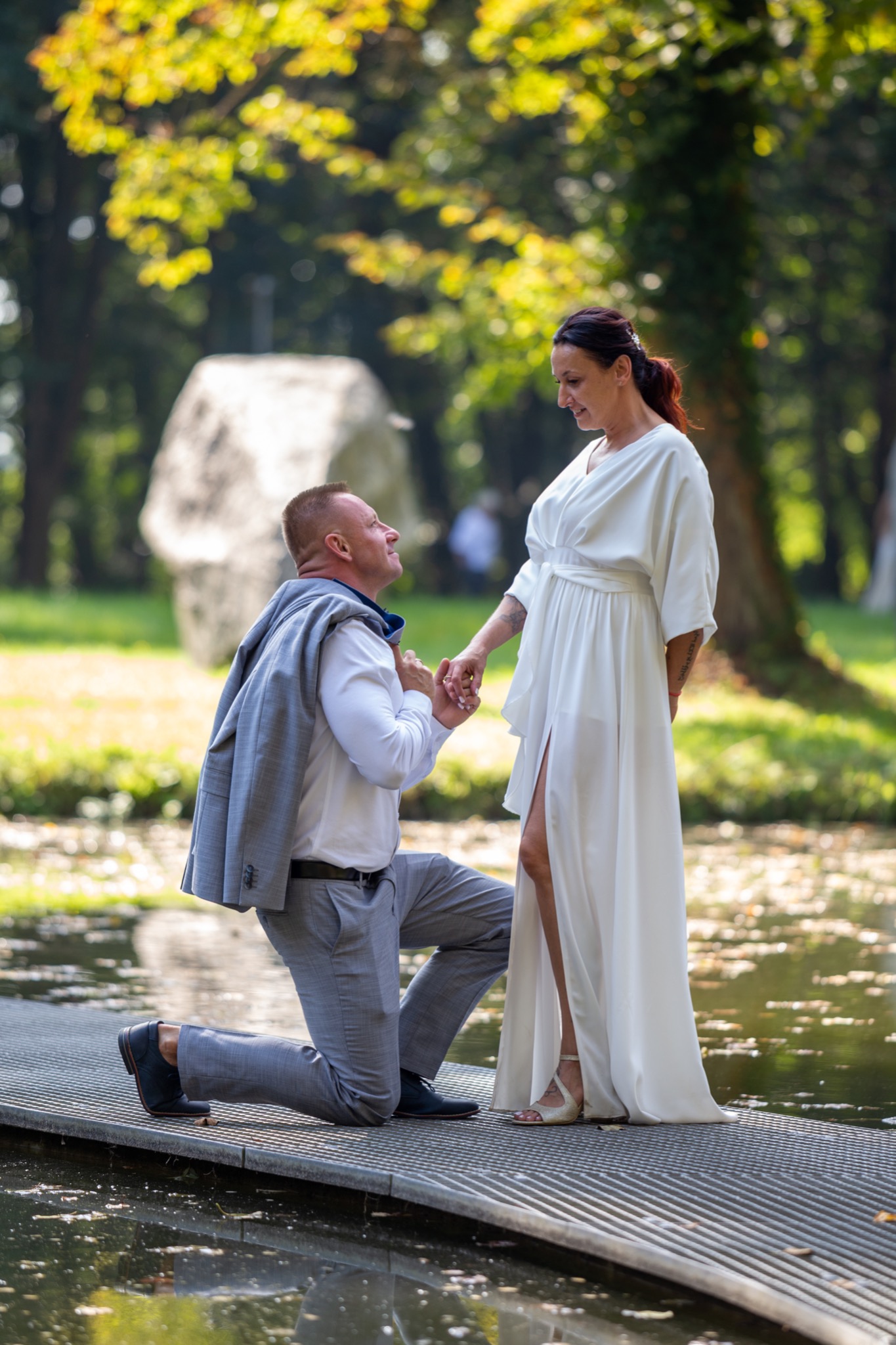 Mężczyzna klęczy na metalowym pomoście nad wodą, trzymając za rękę kobietę w białej sukni, w tle park i duży kamień.