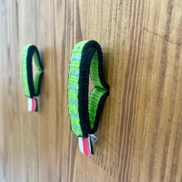 PAPAJ Sp. z o.o. - Dwa uchwyty z zielonej taśmy z czarnym obszyciem i czerwono-białym akcentem, przymocowane do drewnianej powierzchni.