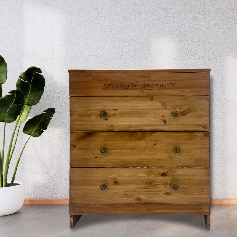 Drewniana komoda z czterema szufladami, ozdobnymi uchwytami w kształcie sterów i wygrawerowanymi współrzędnymi geograficznymi na górnej krawędzi, stojąca na tle białej ściany obok rośliny doniczkowej.