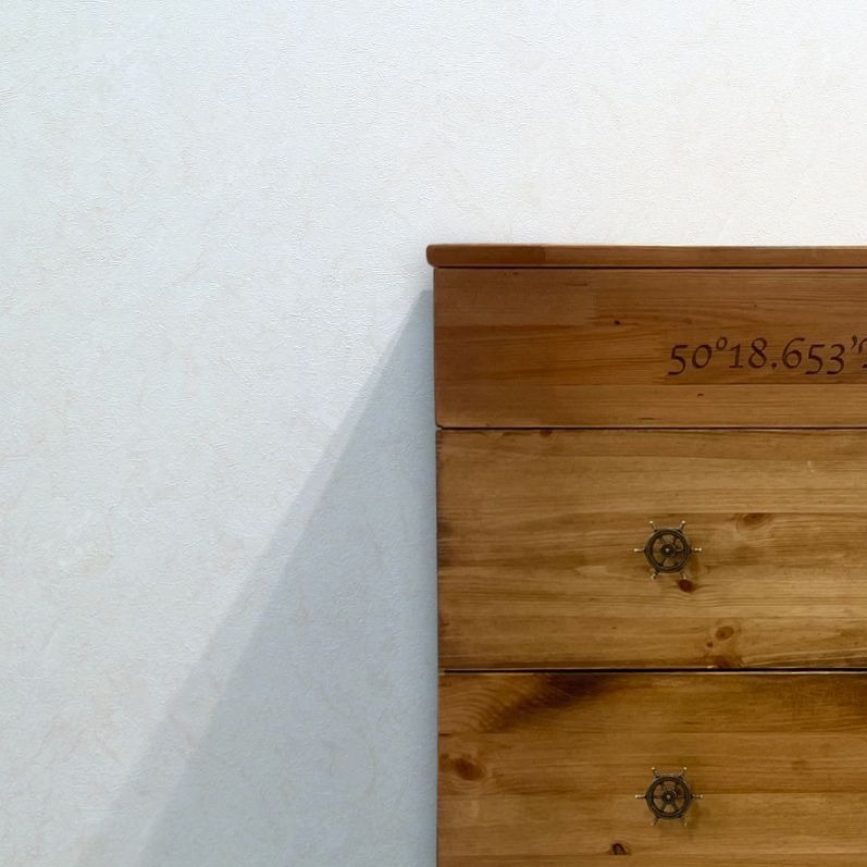 Drewniana komoda z trzema szufladami, z uchwytami w kształcie sterów i wygrawerowanymi współrzędnymi geograficznymi, stojąca przy białej ścianie.