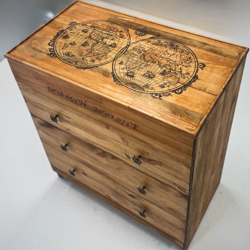 Drewniana komoda z trzema szufladami, zdobiona mapami świata w stylu vintage i współrzędnymi geograficznymi, wykończona w ciepłym odcieniu brązu.