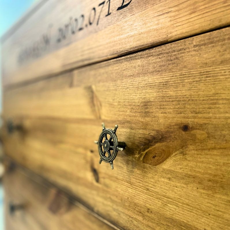 Fragment komody z drewna sosnowego z widocznymi trzema szufladami, każda z uchwytem w kształcie koła sterowego; na górnej szufladzie wygrawerowany napis.