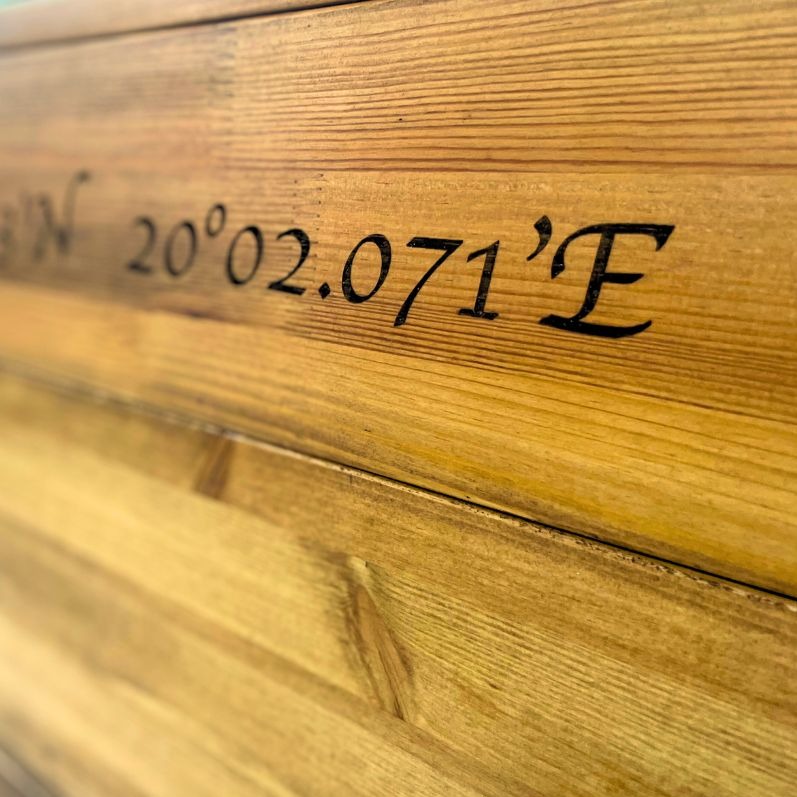 Fragment drewnianej powierzchni z wygrawerowanymi czarnymi współrzędnymi geograficznymi.