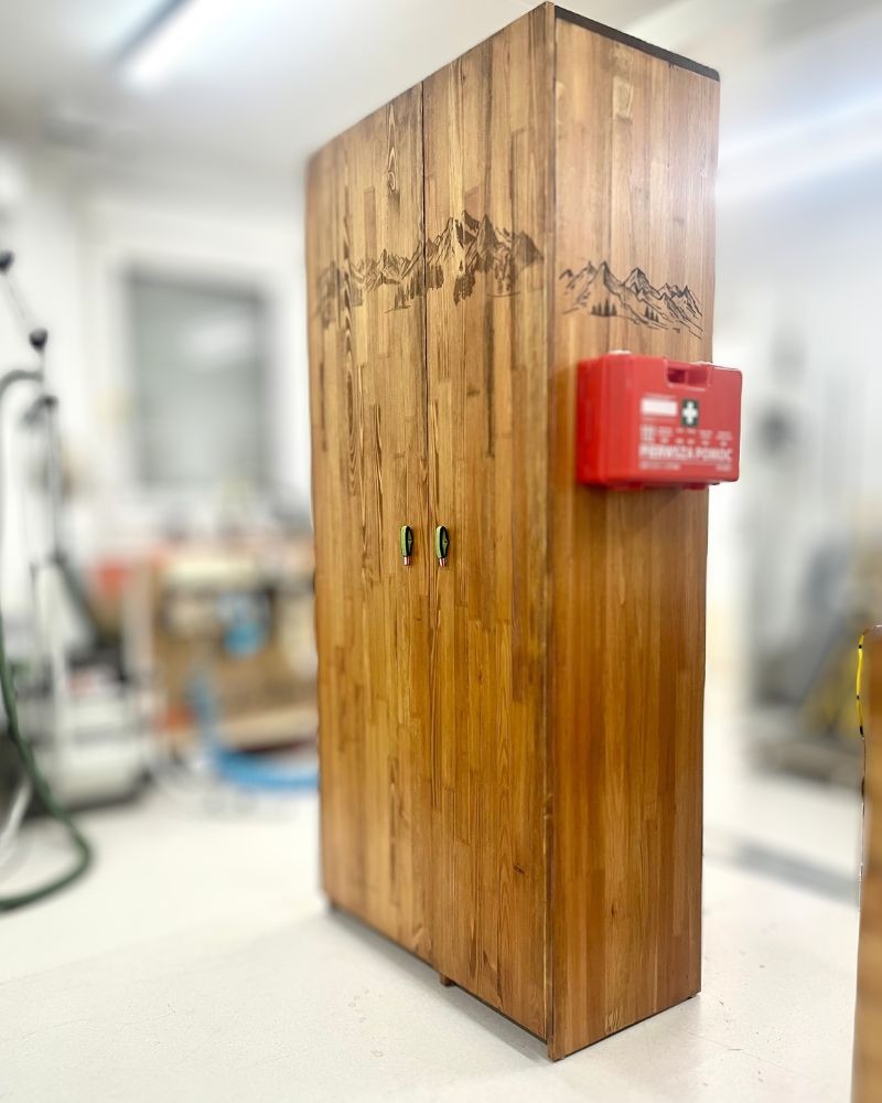Drewniana szafa z motywem górskim i apteczką przymocowaną z boku, widoczna w warsztacie.