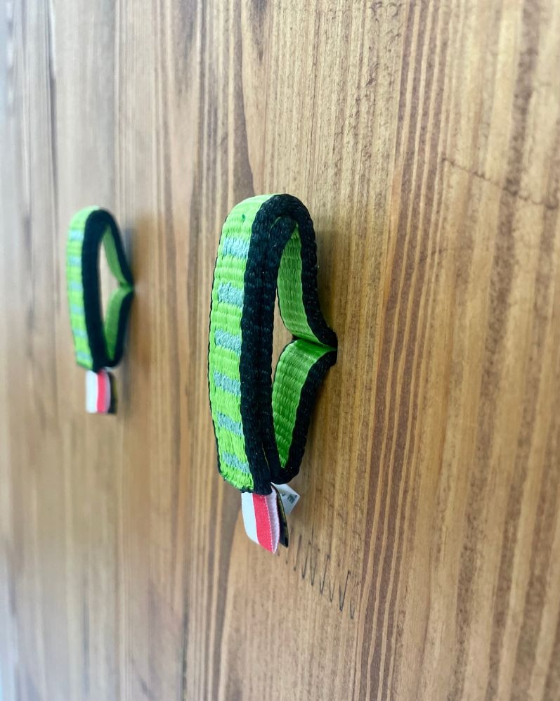 Dwa uchwyty z zielonej taśmy z czarnym obszyciem i czerwono-białym akcentem, przymocowane do drewnianej powierzchni.