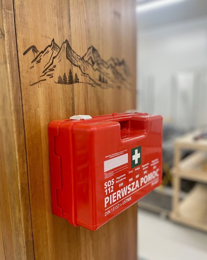 Czerwona apteczka pierwszej pomocy zamontowana na drewnianej szafie z wygrawerowanym motywem górskim.
