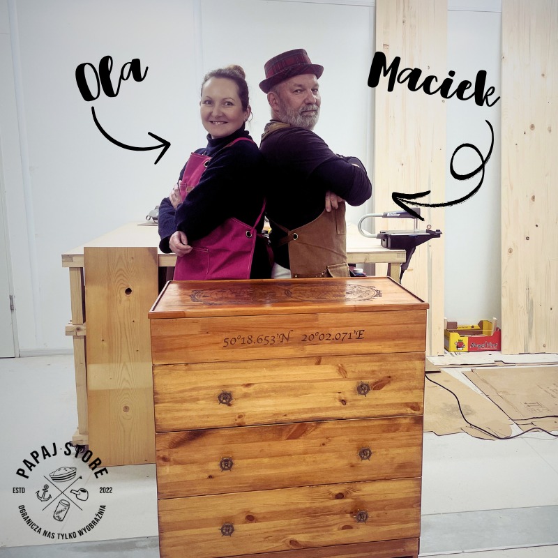Drewniana komoda z czterema szufladami, ozdobiona grawerowanymi współrzędnymi geograficznymi i motywami marynistycznymi, prezentowana przez dwoje stolarzy w fartuchach, stojących tyłem do siebie...