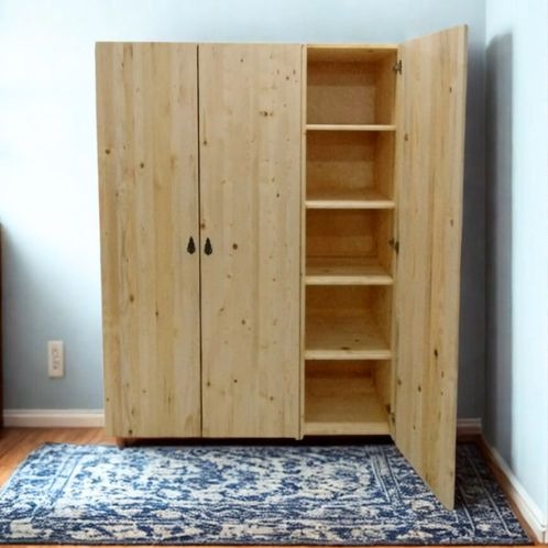 Drewniana szafa z otwartymi drzwiami, ukazująca półki wewnątrz. Widoczny wzór drewna i dywan z niebieskim ornamentem. Prosta forma, naturalne wykończenie.