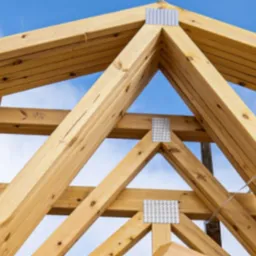 Fragment drewnianej więźby dachowej z metalowymi łącznikami na tle błękitnego nieba. Widoczne detale konstrukcji i łączeń.