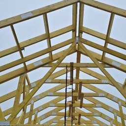 M.A.M. DREWNO MARCIN STACHOWIAK - Wznoszenie konstrukcji drewnianej dachu z widocznymi elementami złącznymi i fragmentem murowanej ściany. Ujęcie z dołu ukazuje etap budowy.