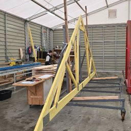 M.A.M. DREWNO MARCIN STACHOWIAK - W hali produkcyjnej: pracownik obsługuje maszynę do cięcia drewna, w tle widoczna konstrukcja więźby dachowej w trakcie montażu i czerwony wózek transportowy.