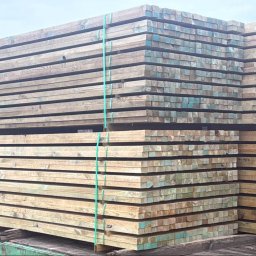 M.A.M. DREWNO MARCIN STACHOWIAK - Stosy impregnowanych desek na paletach, zabezpieczone zielonymi taśmami. Widoczne ślady impregnacji na końcach desek.