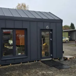 Nowoczesny, grafitowy budynek biurowy z blachy trapezowej, z oknem i drzwiami, na tle placu z zaparkowanymi samochodami. Widoczny fragment ogrodzenia i reklamy.