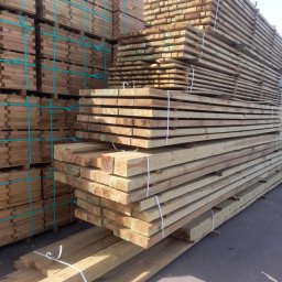 M.A.M. DREWNO MARCIN STACHOWIAK - Stosy tarcicy budowlanej, zabezpieczone taśmami, ułożone na placu składowym pod błękitnym niebem. Widoczne różne przekroje i długości drewna.