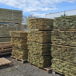 M.A.M. DREWNO MARCIN STACHOWIAK - Stosy impregnowanego drewna budowlanego, ułożone na paletach, widoczne na placu składowym pod błękitnym niebem z chmurami. Drewno zabezpieczone taśmami.