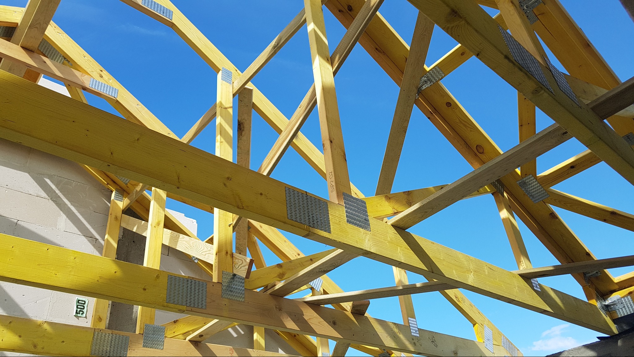 Żółta drewniana więźba dachowa z metalowymi łącznikami na tle błękitnego nieba. Widoczne fragmenty muru z betonu komórkowego.