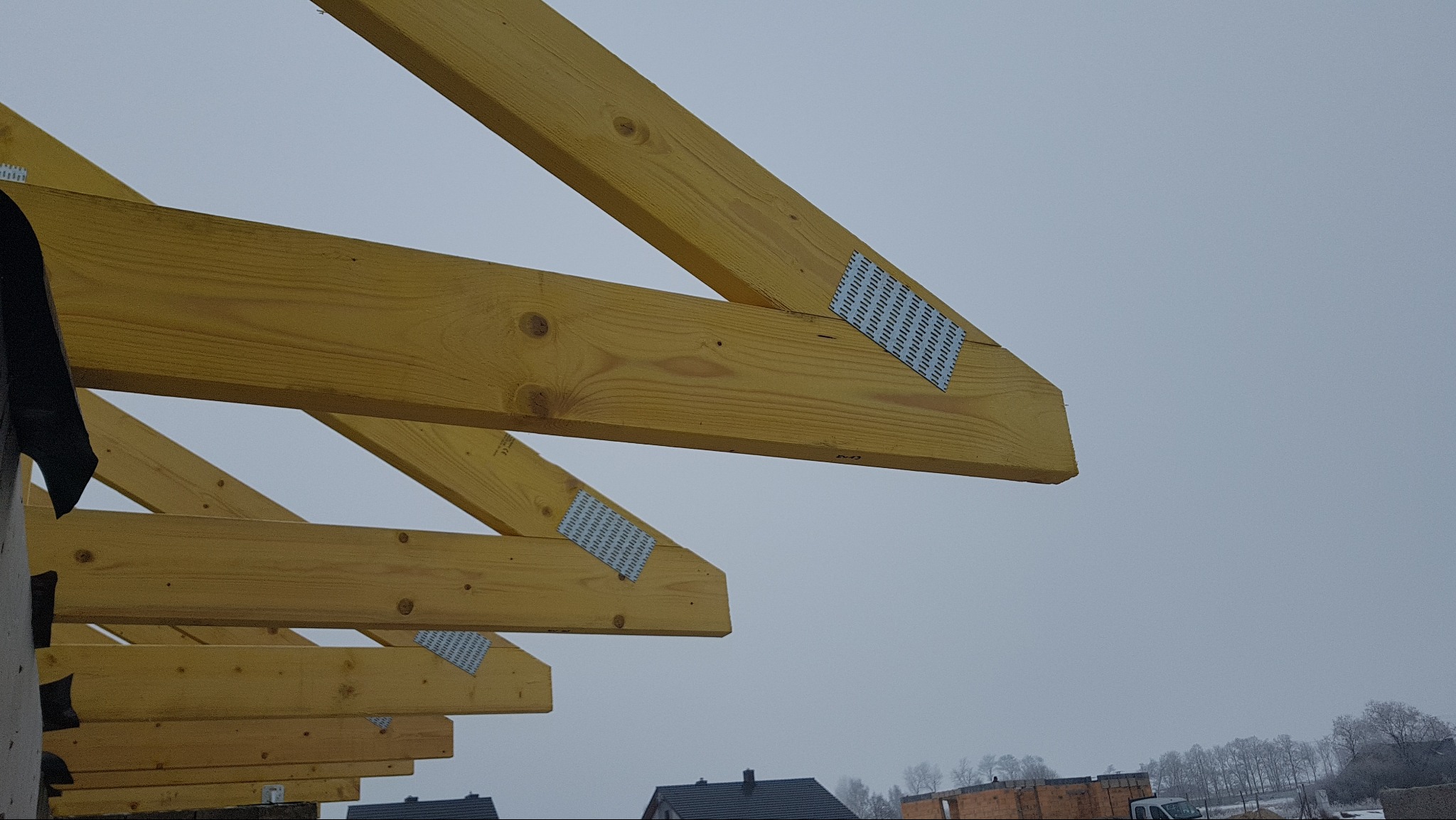 Wiązary dachowe z jasnego drewna, połączone metalowymi płytkami, widoczne na tle szarego nieba. Realizacja z Gniezna.
