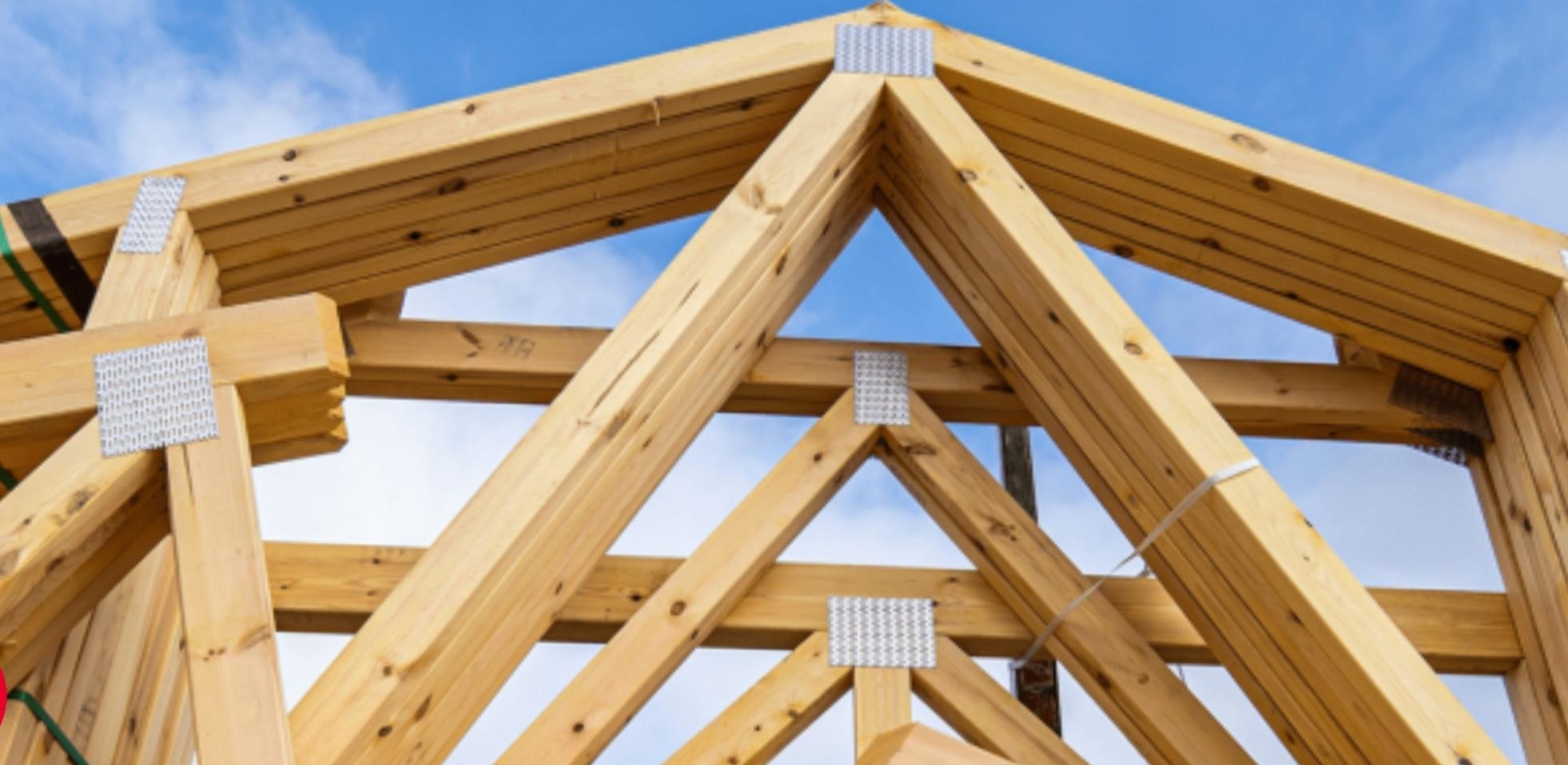 Fragment drewnianej więźby dachowej z metalowymi łącznikami na tle błękitnego nieba. Widoczne detale konstrukcji i łączeń.