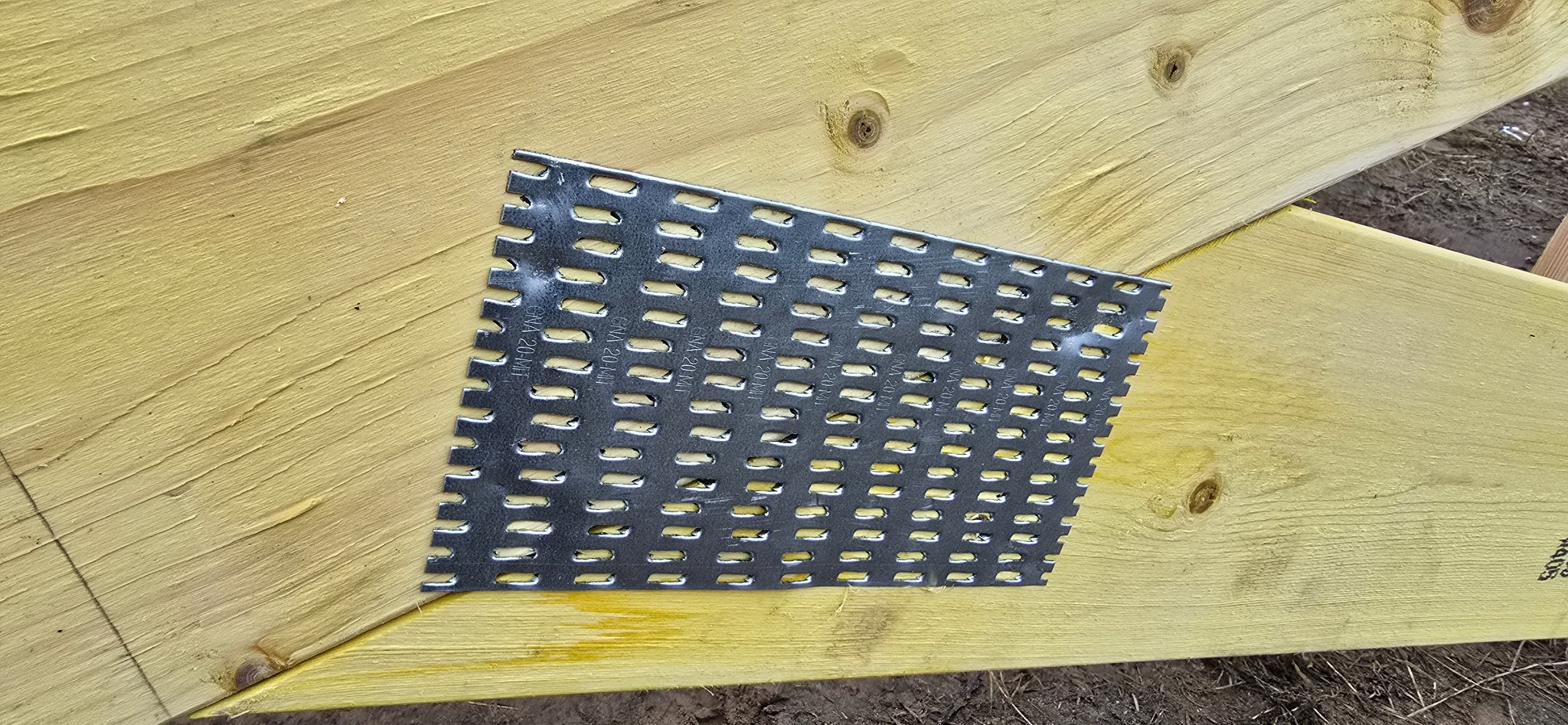 Metalowa płytka perforowana GNA 20-MM łączy dwie drewniane belki w żółtym kolorze. Widoczne słoje drewna i ziemiste tło. Kątowe ujęcie.