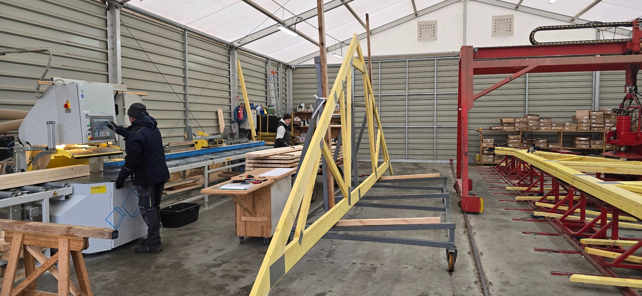 W hali produkcyjnej: pracownik obsługuje maszynę do cięcia drewna, w tle widoczna konstrukcja więźby dachowej w trakcie montażu i czerwony wózek transportowy.